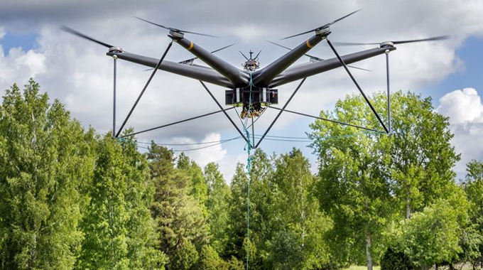 A large, advanced drone equipped with multiple rotors hovers in the air above a lush green forest. 