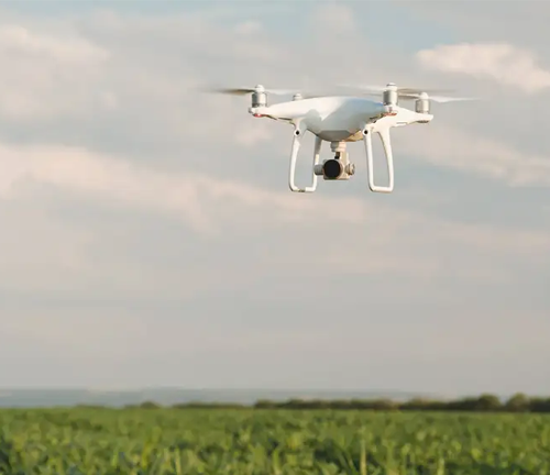 A white drone hovers in the sky above a green field, with a slightly cloudy sky in the background. 