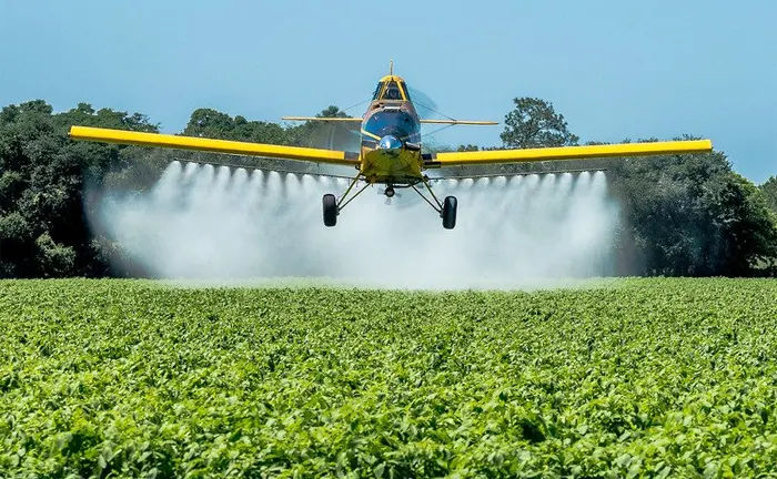 A crop-dusting plane spraying pesticides over a dense green field for pest management.