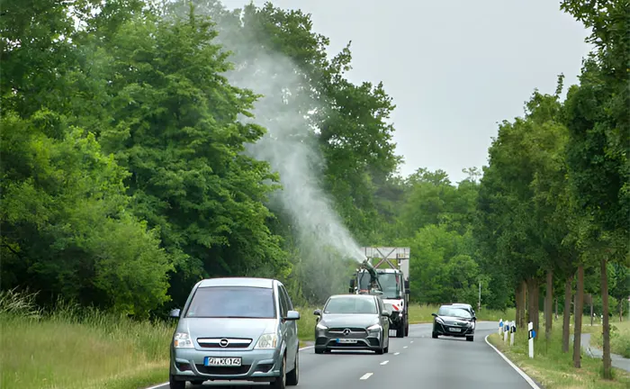 Spraying truck emitting pesticide along a tree-lined road with passing cars.