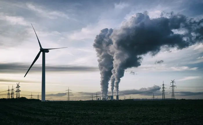 Wind turbine beside power plant emitting smoke, highlighting the contrast between renewable energy and pollution.