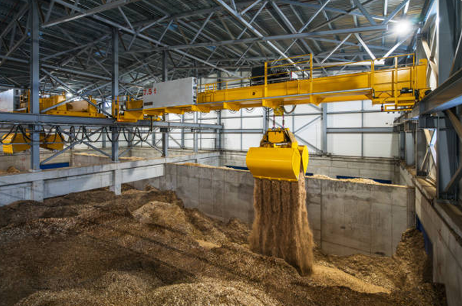 A yellow overhead crane lifts wood chips in a large industrial facility, moving them into a designated compartment. The setup illustrates the biomass handling process for energy production or manufacturing.