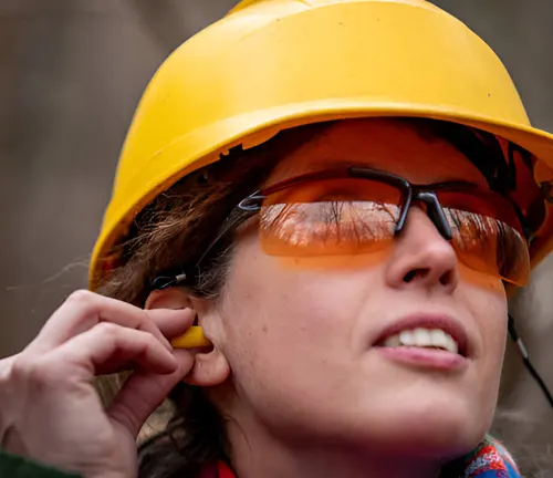 Woman with helmet and safety glasses inserting earplugs for eye and ear protection.