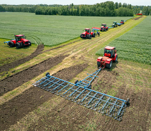 Multiple red tractors operate in a large field, each equipped with different farming implements. The scene shows coordinated agricultural work, with green crops and tilled soil visible.