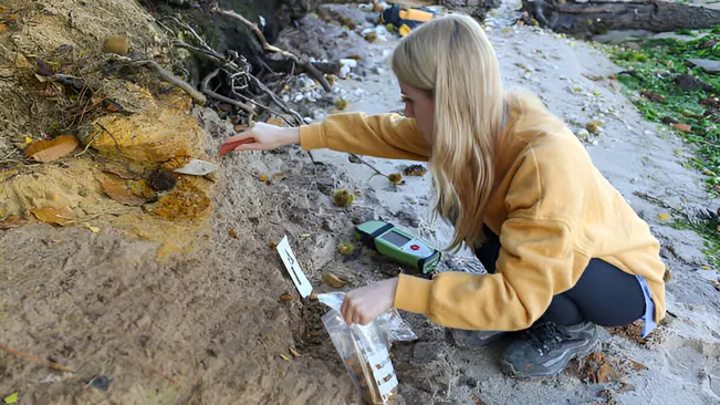 A woman collecting soil samples on a slope for assessing erosion risks.