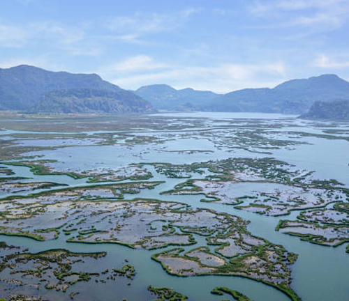 Aerial view of an expansive wetland area with numerous small islands and waterways, surrounded by distant mountain ranges under a blue sky. 