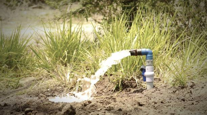 A blue and white PVC pipe spouting a stream of clear water into a small dirt patch, surrounded by tall green grass, indicating an irrigation or groundwater extraction setup in a rural or agricultural area.