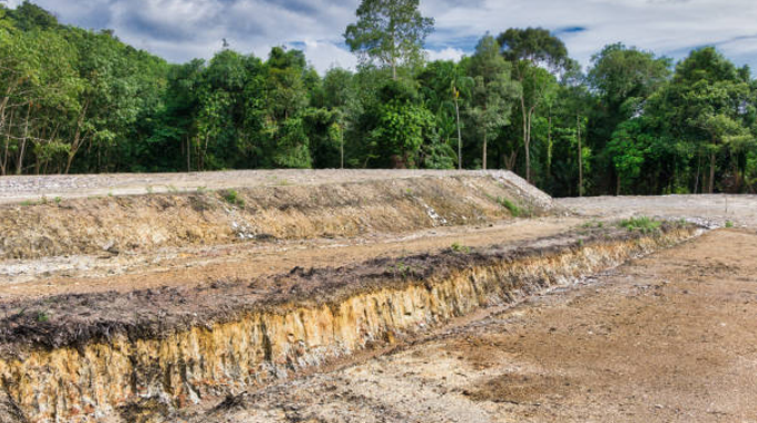 A cleared and exposed patch of earth with visible soil layers, surrounded by a dense green forest, illustrating the impact of deforestation and soil degradation near a forested area.