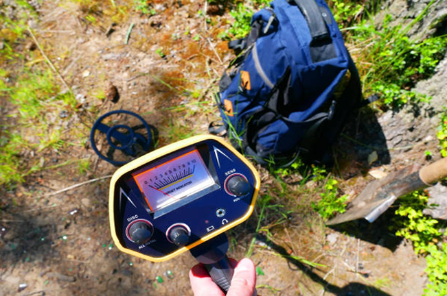 "A hand holds a metal detector displaying a reading, with a backpack and shovel on the ground nearby. The scene suggests outdoor exploration or soil research in a natural setting."