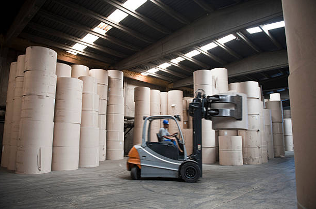 A worker operates a forklift in a large warehouse filled with tall stacks of rolled paper. The scene captures the industrial setting of the pulp and paper industry.