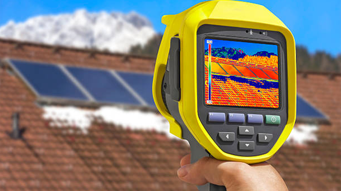 A handheld thermal imaging camera captures a heat map of solar panels on a rooftop, with snow-covered mountains in the background. 