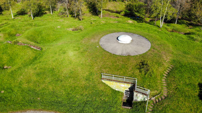 Aerial view of a grassy landscape featuring a circular concrete structure with a dome in the center, and a small fenced platform with steps leading up to it. The area is surrounded by trees and natural vegetation.