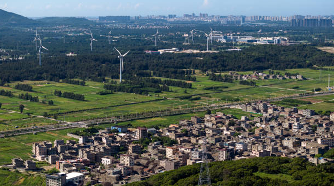 Aerial view of a rural area with a cluster of traditional houses in the foreground, expansive green fields and forests, and several wind turbines scattered across the landscape, with a modern cityscape visible in the distant background.