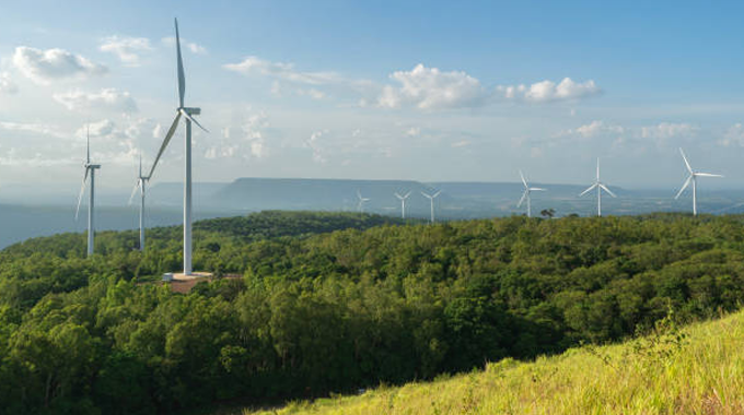 Wind turbines on a lush, green hillside, overlooking a forested landscape with mountains in the background, under a partly cloudy sky.