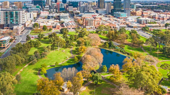 Aerial view of an urban park with lush green trees, a large pond, and surrounding pathways, set against a backdrop of city buildings and infrastructure.