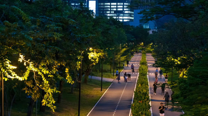 A well-lit urban park pathway at dusk with people walking and cycling, flanked by lush green trees and foliage, with modern buildings illuminated in the background.
