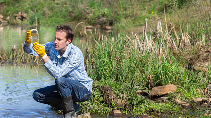 A scientist wearing gloves and rubber boots is crouching by the edge of a pond, holding a water sample container up to the light for inspection. 