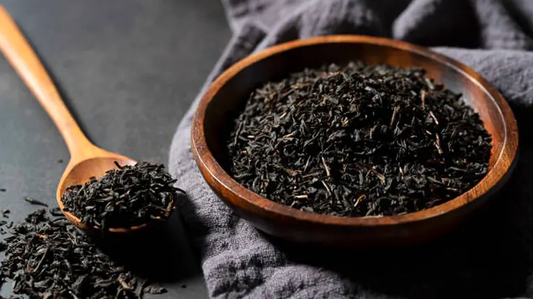 Dried loose tea leaves in a wooden bowl with a spoon on a dark surface, partially covered by a gray cloth.