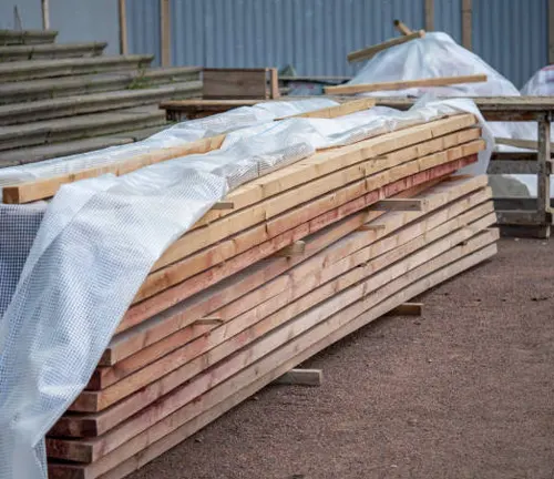 Timber planks covered with protective plastic sheeting, stored outdoors at a construction site.