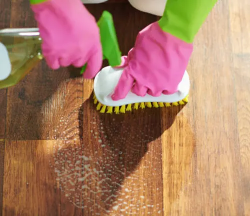 Person scrubbing a wood floor with gloves, brush, and cleaning solution for maintenance.