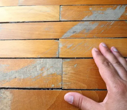 Hand inspecting damaged wood floor with visible cracks and surface imperfections needing repair.