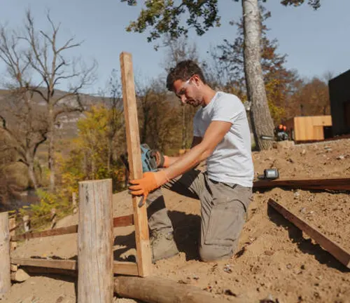 A man installing wooden fence posts outdoors in a sandy, tree-filled landscape area.