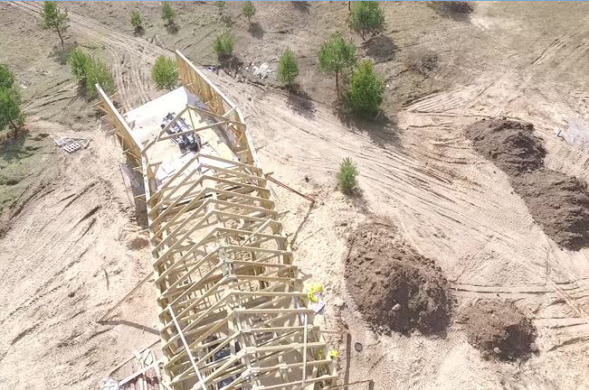 Aerial view of a wooden bridge under construction in a rural area, surrounded by small trees and disturbed soil, illustrating infrastructure development in natural landscapes.