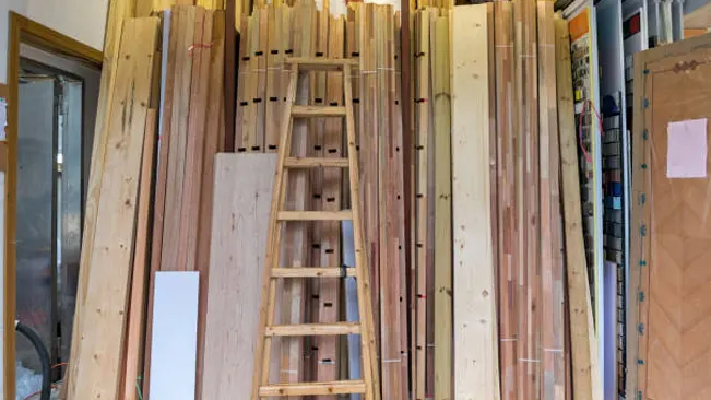 Stacked timber planks stored vertically against a wall with a wooden ladder in between.