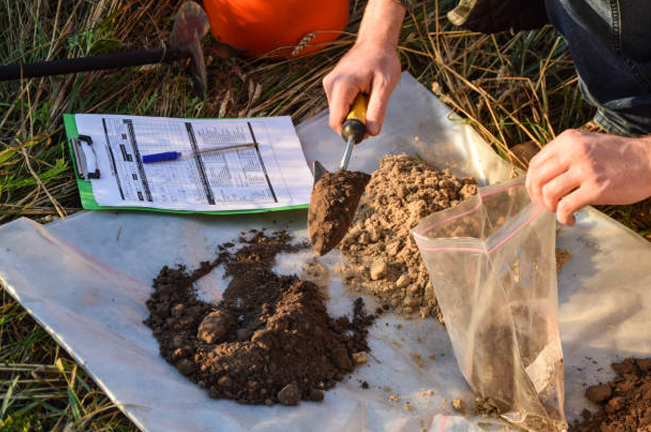 A person collects soil samples with a trowel, placing them into a plastic bag for testing. A clipboard with data sheets and tools lie nearby, indicating soil analysis work.