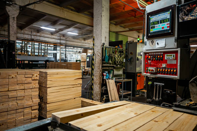 A sawmill's interior with stacks of processed wooden planks beside automated machinery. A digital control panel displays real-time data, reflecting a modern wood production process.