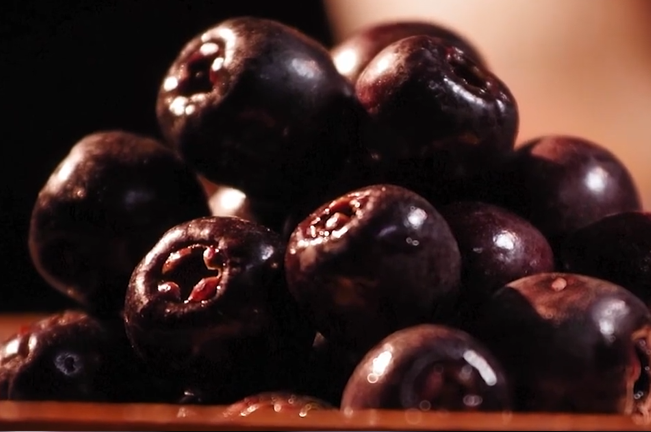 A close-up of a pile of freshly harvested Lipote fruits, showcasing their dark purple, almost black skin. The fruits have a glossy appearance, highlighting their ripeness.