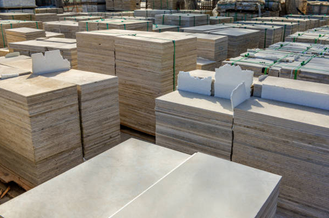 Stacks of stone tiles neatly arranged outdoors in a storage yard. The tiles are in large, uniform bundles, some showing chipped edges, ready for construction or installation.