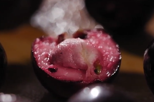 A close-up of a halved Lipote fruit showing its vibrant pink flesh and central seed. The juicy texture and bright color highlight the fruit's tart, sweet interior.