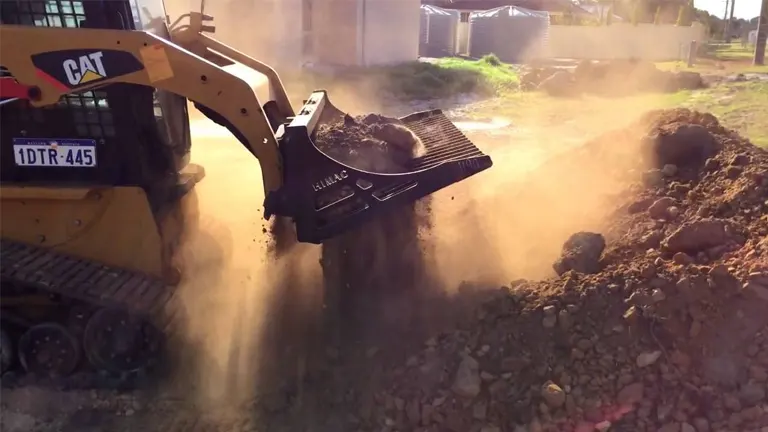 Skid steer using a skeleton bucket to sift and remove soil and rocks during land clearing.