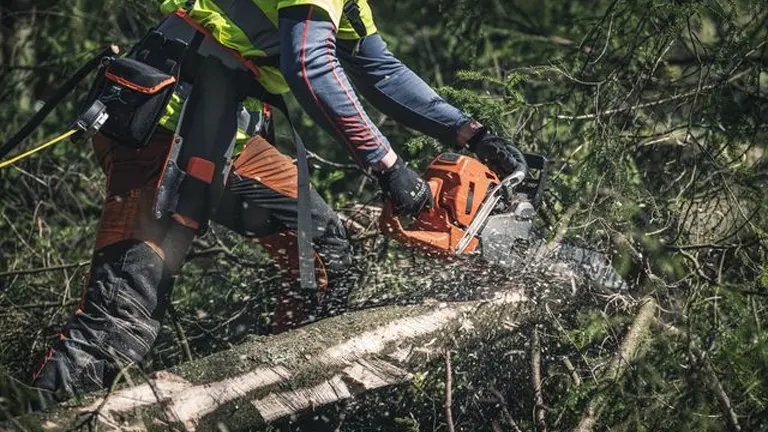 Worker using a chainsaw to safely cut through a large tree branch during storm cleanup, wearing full protective gear including gloves, helmet, and safety pants.