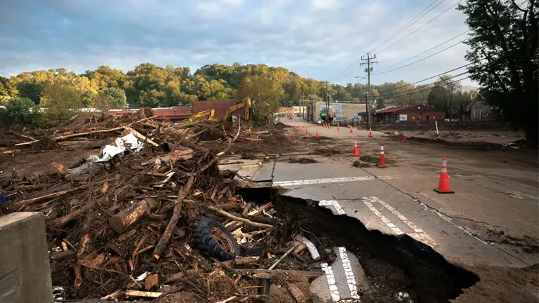 Severe hurricane damage in a town, with debris and fallen trees scattered, a large crack in the road, and cleanup efforts underway as part of recovery.