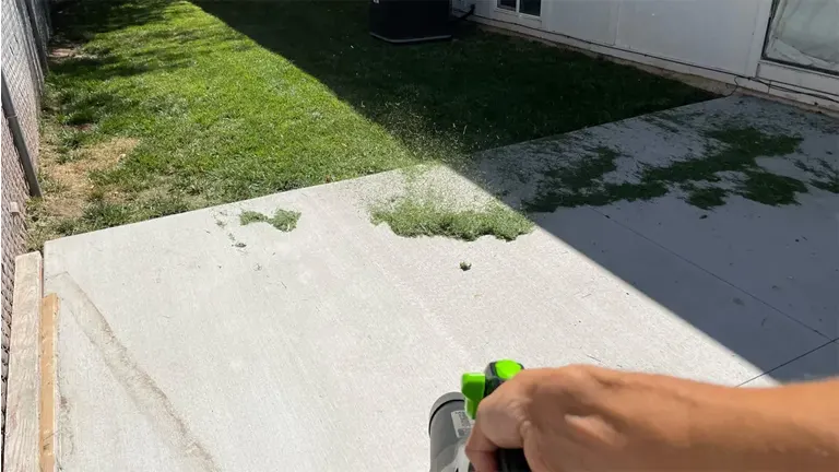 Hand using the Greenworks Pro 80V leaf blower to clear grass clippings from a concrete patio next to a lawn.